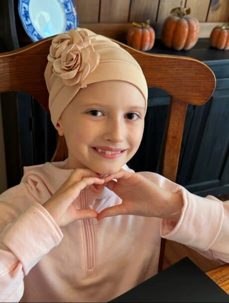 A smiling child wearing a beige headscarf and making a heart shape with hands.