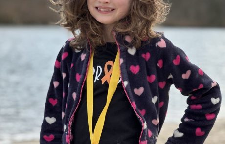 Young girl wearing a heart-patterned jacket and a gold medal, smiling outdoors.