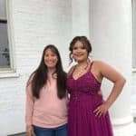Two young women posing together, one in a pink sweater and the other in a purple formal dress.