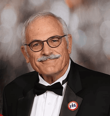 Elderly man in a tuxedo with glasses and a pin smiles warmly.