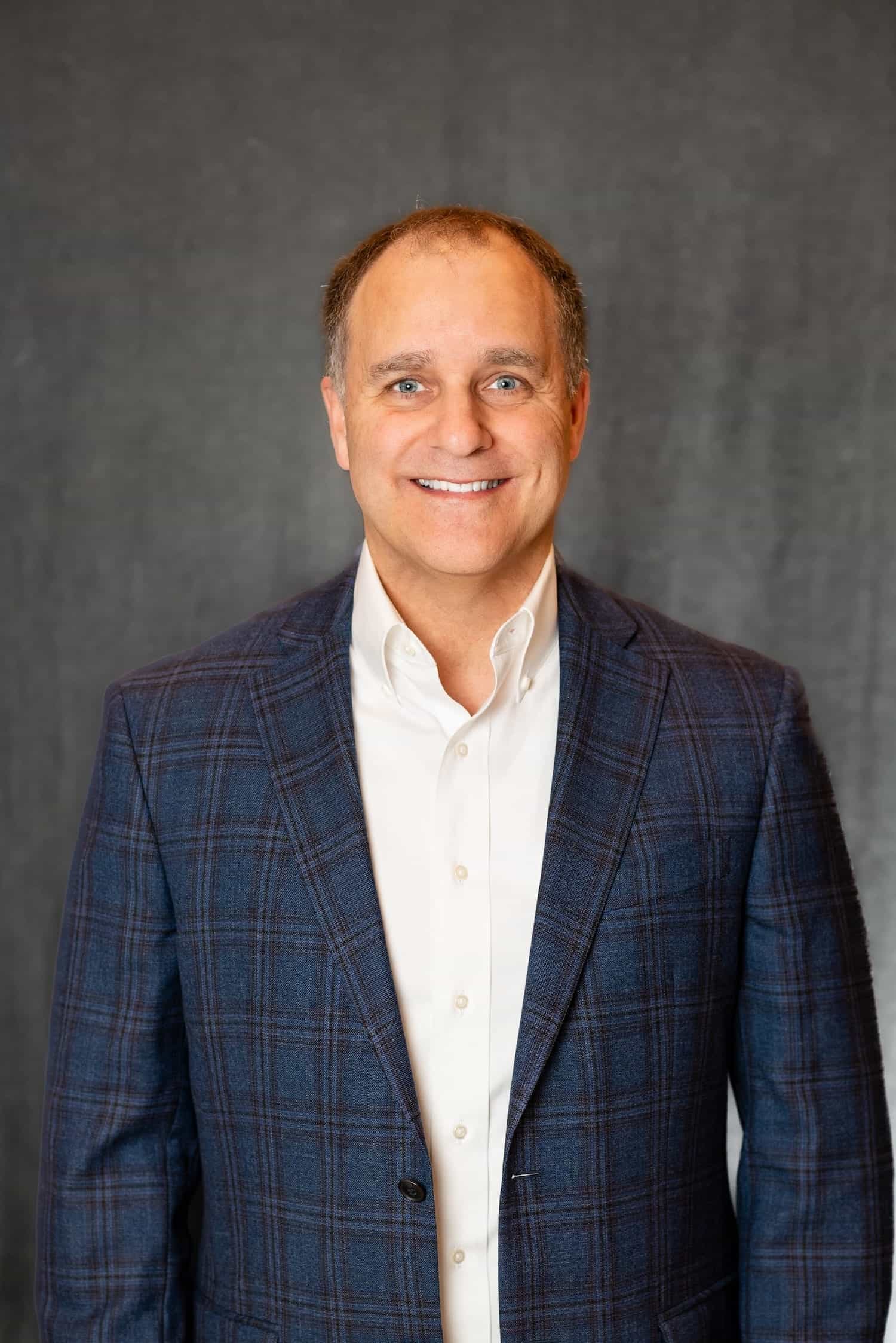 Smiling man in a blue checkered blazer and white shirt against a grey background.