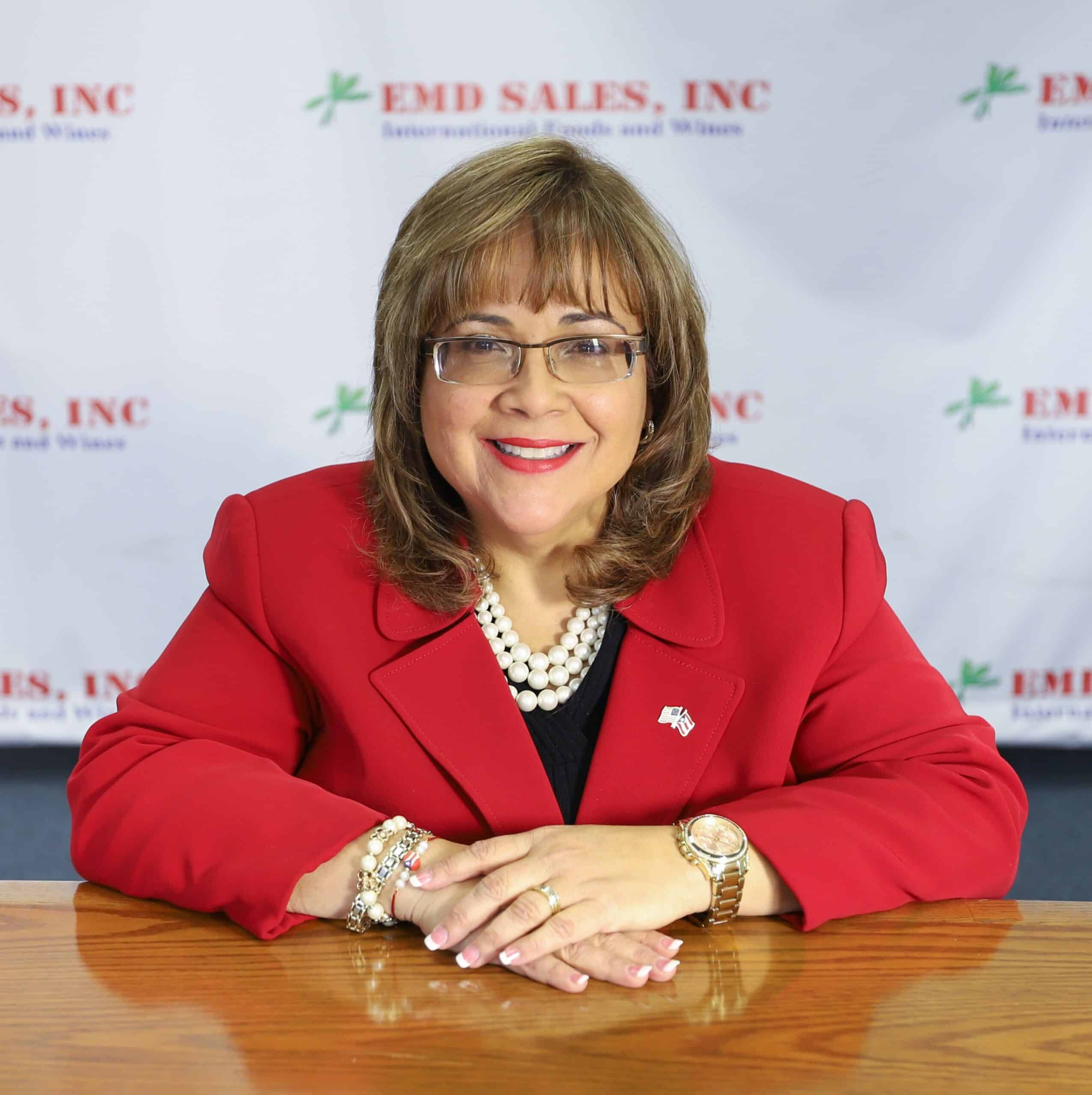 Woman in red blazer smiling at a desk with a branded backdrop.