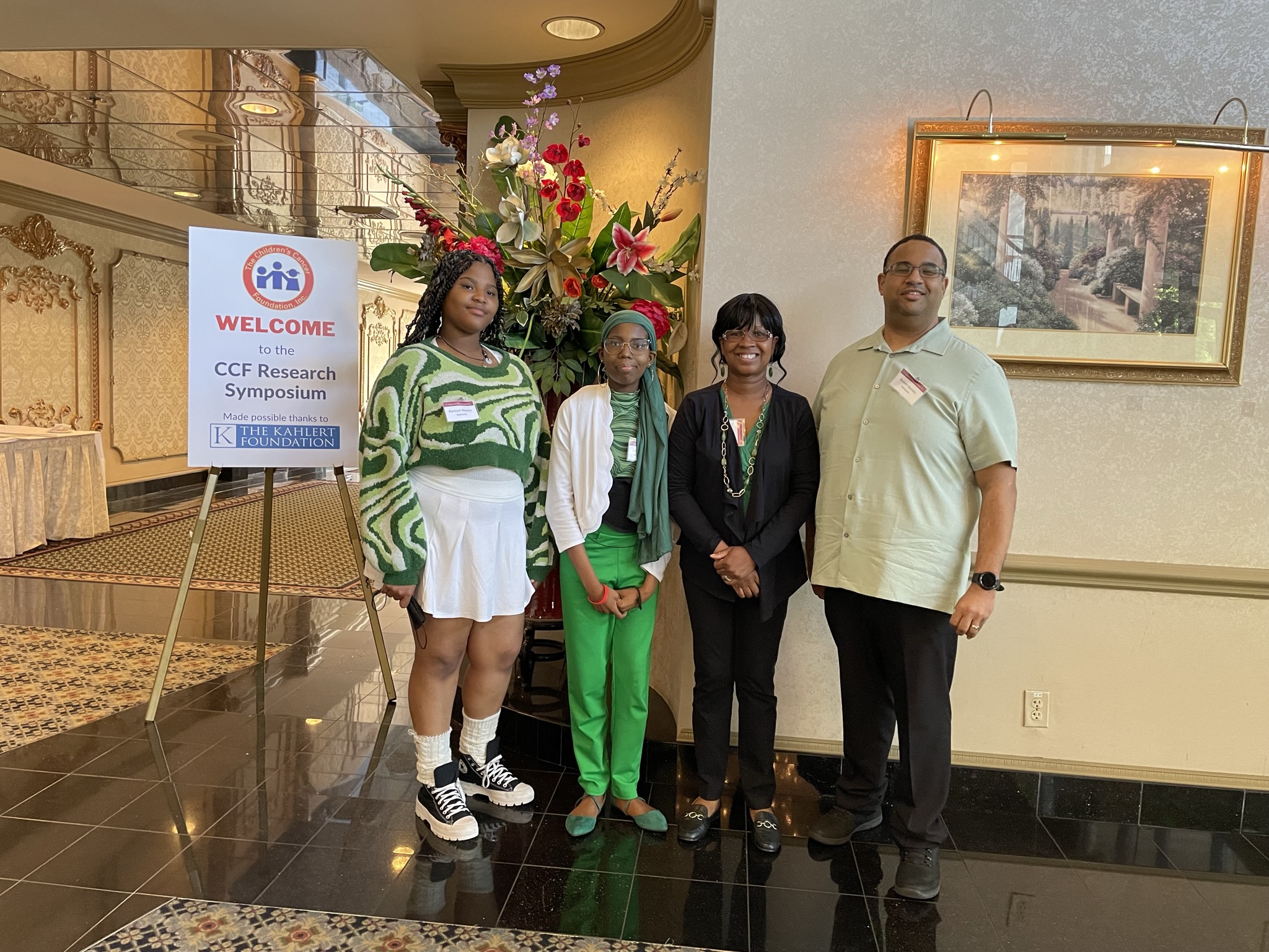 Four people posing indoors near a floral arrangement and a sign.